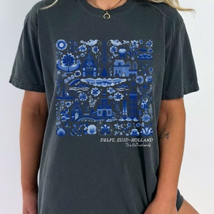 K&ouml;nnte beinhalten: Ein dunkelgraues T-Shirt mit einem blauen und wei&szlig;en Grafikdruck einer holl&auml;ndischen Dorfszene. Die Grafik enth&auml;lt H&auml;user, Blumen und Boote. Der Text "DELFT, ZUID-HOLLAND The Netherlands" ist unterhalb der Grafik gedruckt.