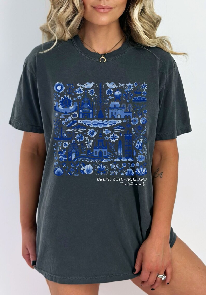 K&ouml;nnte beinhalten: Ein dunkelgraues T-Shirt mit einem blauen und wei&szlig;en Grafikdruck einer holl&auml;ndischen Dorfszene. Die Grafik enth&auml;lt H&auml;user, Blumen und Boote. Der Text "DELFT, ZUID-HOLLAND The Netherlands" ist unterhalb der Grafik gedruckt.