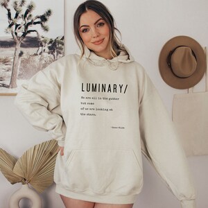May include: A cream-colored hoodie with a black print that reads "LUMINARY/ We are all in the gutter but some of us are looking at the stars. - Oscar Wilde"