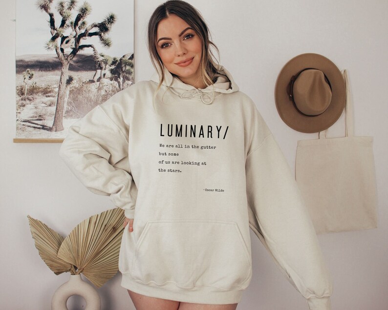 May include: A cream-colored hoodie with a black print that reads "LUMINARY/ We are all in the gutter but some of us are looking at the stars. - Oscar Wilde"