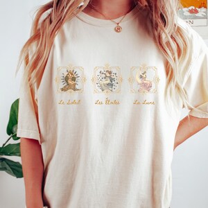 May include: A cream-colored t-shirt with a graphic print of three celestial figures in gold frames. The text "Le Soleil", "Les &Eacute;toiles", and "La Lune" is printed below each figure.