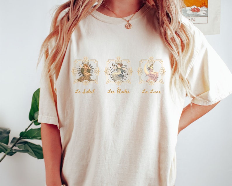 May include: A cream-colored t-shirt with a graphic print of three celestial figures in gold frames. The text "Le Soleil", "Les &Eacute;toiles", and "La Lune" is printed below each figure.