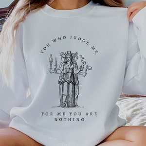 Op de afbeelding: Witte trui met een zwarte grafische print van een driekoppige figuur met de tekst "You who judge me for me you are nothing."