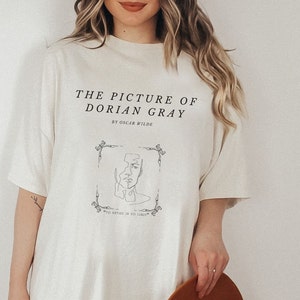 May include: A white t-shirt with a black graphic print of a book cover. The text on the shirt reads "The Picture of Dorian Gray" by Oscar Wilde. The quote "To define is to limit" is also printed on the shirt.