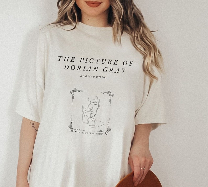 May include: A white t-shirt with a black graphic print of a book cover. The text on the shirt reads "The Picture of Dorian Gray" by Oscar Wilde. The quote "To define is to limit" is also printed on the shirt.