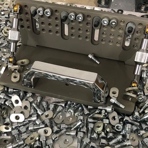 May include: A grey metal jig with two adjustable arms and a chrome handle. The jig is surrounded by a large quantity of silver screws and washers.