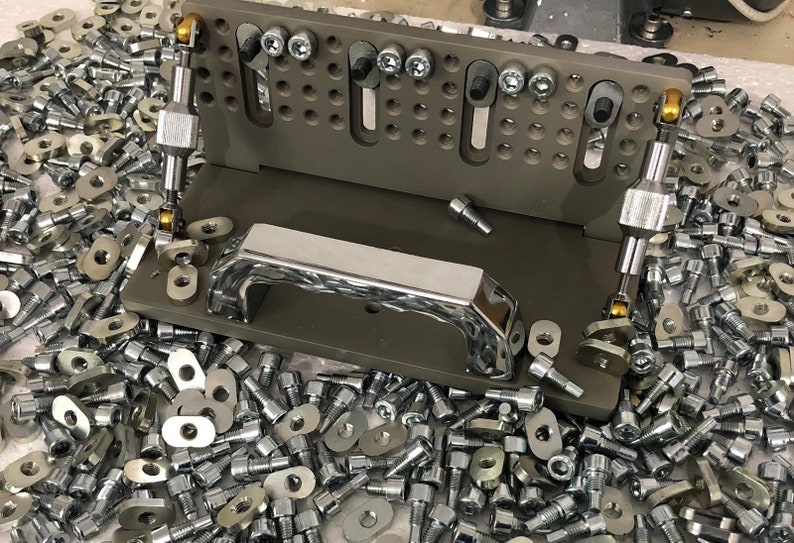 May include: A grey metal jig with two adjustable arms and a chrome handle. The jig is surrounded by a large quantity of silver screws and washers.