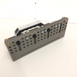 May include: A grey metal workbench with a handle and multiple holes for attaching tools. The workbench has a silver metal handle and black screws.