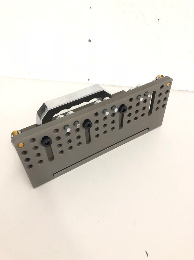 May include: A grey metal workbench with a handle and multiple holes for attaching tools. The workbench has a silver metal handle and black screws.