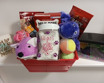 Op de afbeelding: Een rode mand gevuld met hondensnoepjes, speelgoed en andere hondenartikelen. De mand bevat een glazen pot met het label "Ginger's Dog Treats", een paars pluche hondenspeelgoed, een roze touwspeelgoed, een blauw pluche speelgoed en een tennisbal.