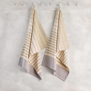 May include: Two beige and gray striped kitchen towels hanging on white hooks. The towels have a striped pattern with a gray border at the bottom. The background is a textured white wall.