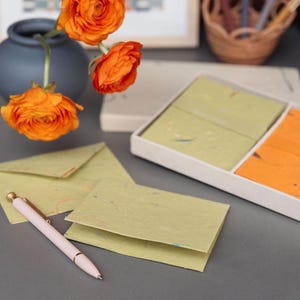Könnte beinhalten: Ein Stillleben mit Schreibwaren, darunter ein Stift, Umschläge und Notizkarten in Grün- und Orangetönen. Eine Vase mit orangefarbenen Blumen und eine dekorative Box sind ebenfalls vorhanden. Auf einer grauen Oberfläche.