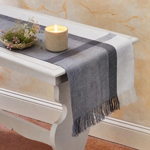 May include: A rectangular table runner featuring a gradient of gray and white tones with a woven texture and fringed ends. A cylindrical beige candle with a lit flame and a small ceramic bowl containing delicate pink and white flowers sit on top.