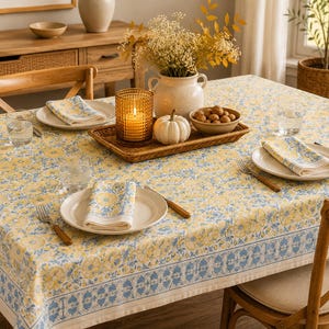 Könnte beinhalten: Eine rechteckige Tischdecke mit gelbem und blauem Blumenmuster und einer dekorativen Bordüre. Der Tisch ist mit weißen Tellern, gefalteten Servietten, Gabeln und Messern gedeckt. Ein Weidentablett enthält eine brennende bernsteinfarbene Kerze.