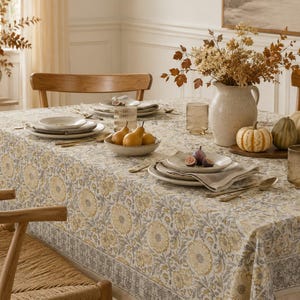 May include: A rectangular tablecloth featuring a repeating floral pattern in muted yellow and grey tones. The fabric has a wide decorative border along the edges and is draped over a dining table set with white plates, glassware, and seasonal decor.