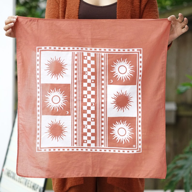 Puede incluir: Un pa&ntilde;uelo de color terracota con un dise&ntilde;o blanco que presenta s&iacute;mbolos repetidos de sol y luna, un patr&oacute;n a cuadros y un borde de cuadrados peque&ntilde;os.