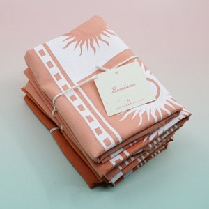 Puede incluir: Una pila de bandanas de color melocot&oacute;n con un dise&ntilde;o de sol blanco y el texto "Bandana by November Fields".