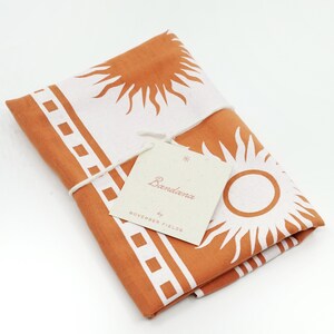 Puede incluir: Un pa&ntilde;uelo naranja doblado con un dise&ntilde;o de sol blanco y un borde blanco. El pa&ntilde;uelo est&aacute; etiquetado como "Bandana by November Fields".