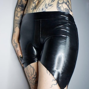 May include: Black latex shorts with a high waist and a unique asymmetrical hemline. The shorts have a glossy finish and a form-fitting design. The shorts are made of a shiny, smooth material.