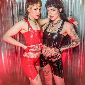 May include: Two people wearing red and black latex outfits pose in front of a metallic background. The red outfit includes a halter top, shorts, and wrist cuffs. The black outfit features a harness top and shorts. Both outfits have multiple straps and buckles.