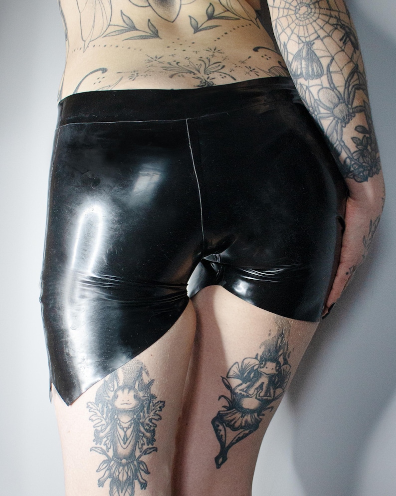 May include: Black latex shorts with a high waist and a glossy finish. The shorts have a unique cut on the leg openings. The image shows the shorts from the back and side.