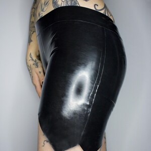 May include: Black latex shorts with a high waistband. The shorts have a shiny finish and a unique cut at the hemline. The garment is form-fitting and appears to be made of a smooth, flexible material. The shorts are designed for a close fit.