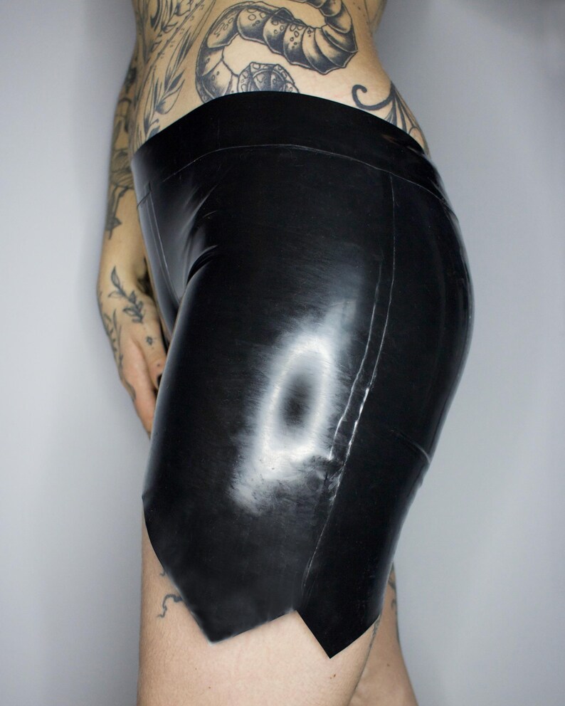 May include: Black latex shorts with a high waistband. The shorts have a shiny finish and a unique cut at the hemline. The garment is form-fitting and appears to be made of a smooth, flexible material. The shorts are designed for a close fit.