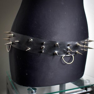 Può includere: Una cintura in pelle nera con punte argentate e un anello in metallo dorato.