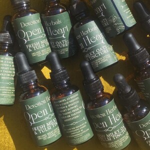 K&ouml;nnte beinhalten: Eine Sammlung kleiner, brauner Glasflaschen mit schwarzen Tropfern. Jede Flasche hat ein gr&uuml;nes Etikett mit wei&szlig;em Text, auf dem "Devotion Herbals Open Heart Fresh Milky & Tulsi Tincture" steht. Die Flaschen sind auf einer gelben Oberfl&auml;che angeordnet.