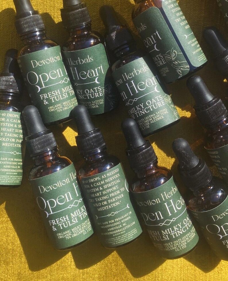 K&ouml;nnte beinhalten: Eine Sammlung kleiner, brauner Glasflaschen mit schwarzen Tropfern. Jede Flasche hat ein gr&uuml;nes Etikett mit wei&szlig;em Text, auf dem "Devotion Herbals Open Heart Fresh Milky & Tulsi Tincture" steht. Die Flaschen sind auf einer gelben Oberfl&auml;che angeordnet.