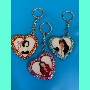 May include: Three heart-shaped keychains with glitter embedded in the resin. Each keychain features a different image of Selena. One keychain has a red glitter heart with a black and white image of Selena. Another keychain has a blue glitter heart with a color image of Selena. The third keychain has a pink glitter heart with a color image of Selena.