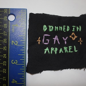 May include: A black fabric square with embroidered text that reads "DONNED IN GAY APPAREL" in green and purple thread. A blue ruler with inch measurements is to the left of the fabric.