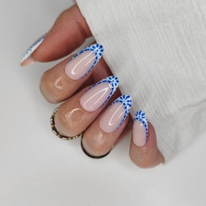May include: Long, oval-shaped fingernails with a light pink base and a blue floral design along the edges. The nails are well-manicured and appear glossy. A gold ring is visible on one finger.
