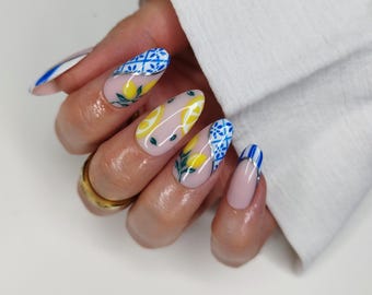 May include: Almond-shaped nails featuring a nude base with intricate blue and white tile patterns, yellow lemon illustrations with green leaves, and silver metallic accents. The design includes blue and white striped tips on some nails.