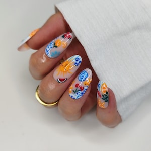 May include: A hand with long, oval-shaped nails, each adorned with colorful, detailed nail art. Designs include a sun, watermelon, cherries, and floral patterns in blue, orange, and red. A gold ring is visible on the ring finger.