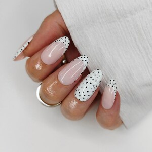 May include: Close-up of a hand displaying long, oval-shaped nails. Some nails are white with black polka dots, others have a pale pink base with a white tip. The nails are well-manicured and have a glossy finish.
