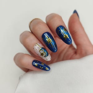 May include: A hand with long, oval-shaped nails. The nails are painted in a celestial theme, featuring a mix of deep blue and off-white colors. Gold stars and crescent moons adorn the nails, creating a starry night effect. The nails are well-manicured and polished.