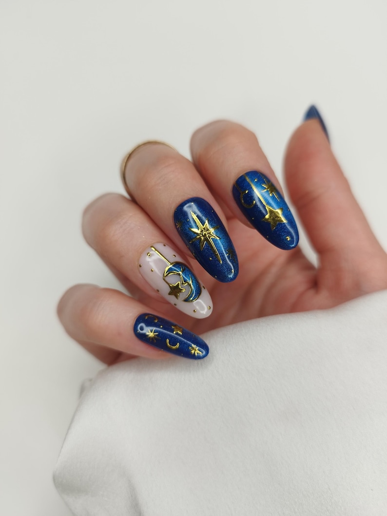 May include: A hand with long, oval-shaped nails. The nails are painted in a celestial theme, featuring a mix of deep blue and off-white colors. Gold stars and crescent moons adorn the nails, creating a starry night effect. The nails are well-manicured and polished.