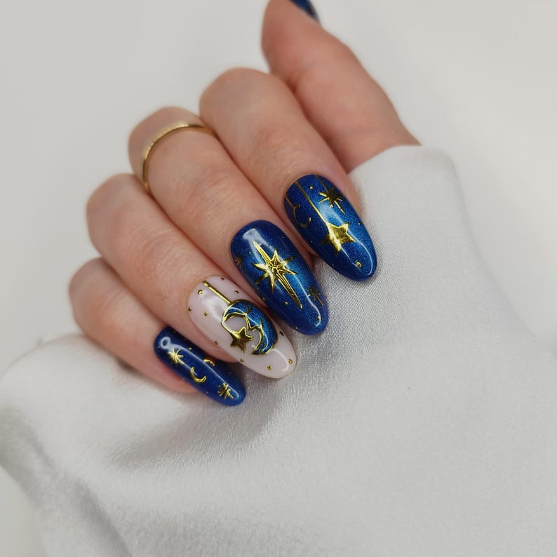 May include: A hand with long, oval-shaped nails. The nails are painted in a celestial theme, with a mix of deep blue and nude colors. Gold star and moon designs adorn the nails, creating a starry night effect. A gold ring is on the ring finger.