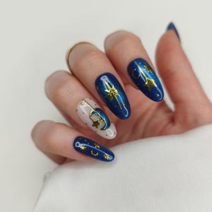 May include: A hand displaying a set of long, oval-shaped nails. The nails are painted in a deep blue and white color scheme, adorned with gold celestial designs including stars and crescent moons. The design is intricate and detailed, creating a celestial theme.
