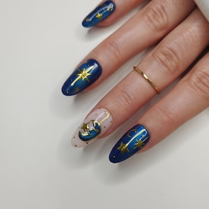 May include: Oval-shaped, long fingernails painted with a celestial theme. The nails feature a dark blue base with gold star and moon designs. One nail has a light pink base with a gold moon and star design. A gold ring is on one finger.