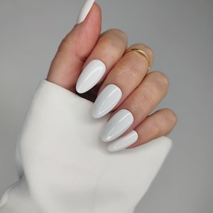 Puede incluir: Primer plano de una mano con cinco uñas blancas ovaladas. Las uñas están pintadas de blanco brillante.