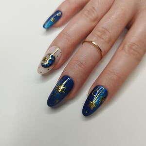 May include: A hand with long, oval-shaped nails, each painted with a celestial theme. The nails feature a combination of blue, gold, and nude colors, with designs including stars, moons, and dots. A gold ring is worn on one finger.