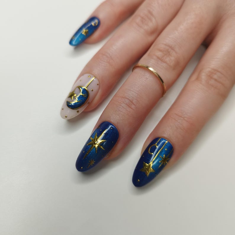 May include: A hand with long, oval-shaped nails, each painted with a celestial theme. The nails feature a combination of blue, gold, and nude colors, with designs including stars, moons, and dots. A gold ring is worn on one finger.