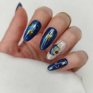 May include: Oval-shaped, dark blue nail art with gold star and crescent moon designs. One nail is light pink with gold dots and a gold moon and star design. The nails have a glossy finish.