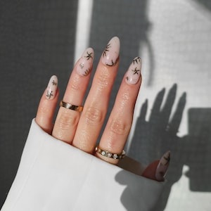 May include: A hand with long, almond-shaped nails painted in a light pink hue. The nails feature a design of gold starfish and gold accents. Two gold rings adorn the fingers, one plain and one with small, clear stones.