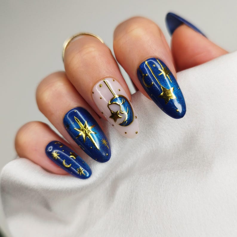 May include: A hand displays long, almond-shaped nails with a celestial theme. The nails feature a deep blue shimmer with gold accents, including stars, moons, and a hanging crescent moon design. One nail is a nude color with gold dots.