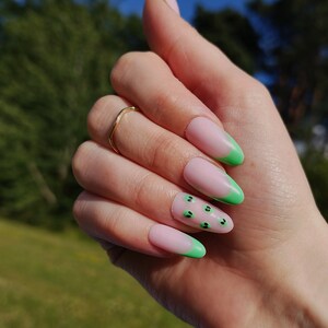 May include: Close-up of a hand with long, almond-shaped nails painted with a light pink base and green tips. The green tips have a small black design on them.