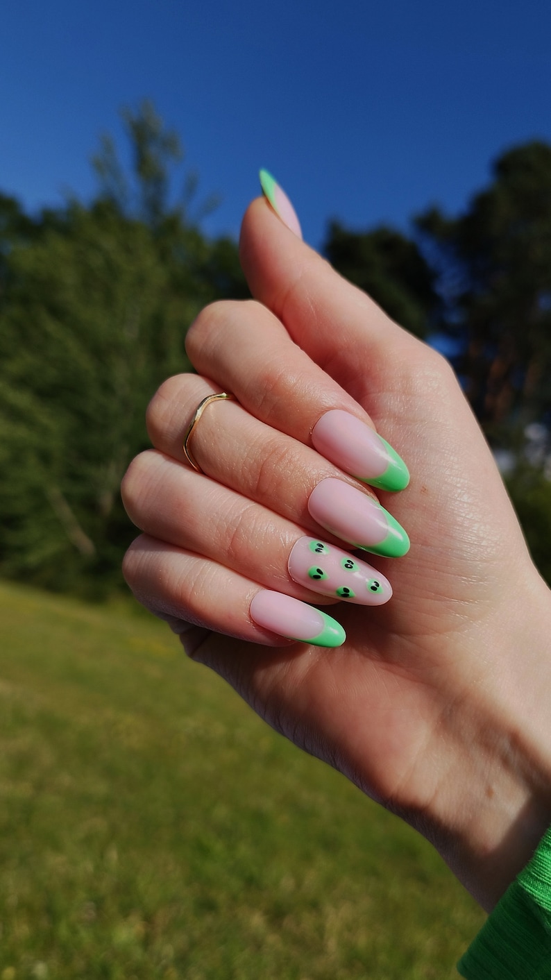May include: Close-up of a hand with long, almond-shaped nails painted with a light pink base and green tips. The green tips have a small black design on them.