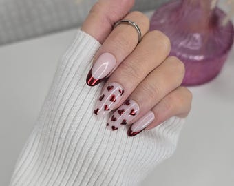 May include: Close-up of a hand with oval-shaped nails painted with a light pink base and a red tip. The nails are decorated with small red glitter hearts.